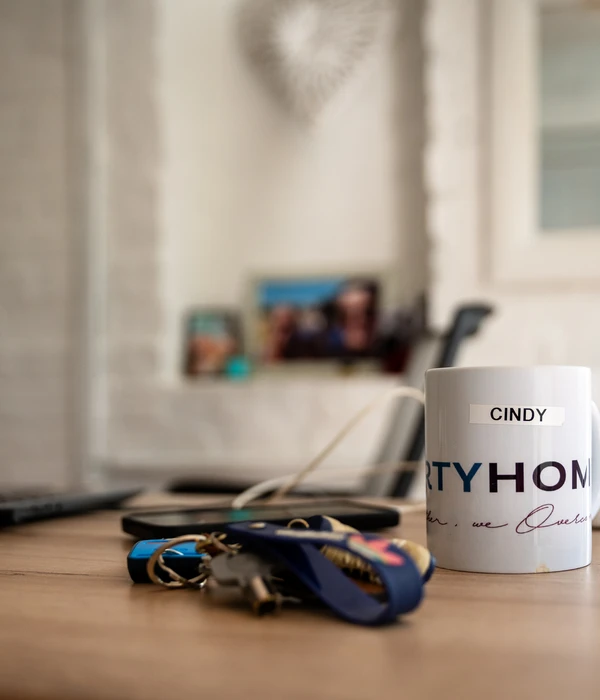 A white mug labeled "Cindy" sits on a wooden desk next to a set of keys, with a blurred background of a home office.