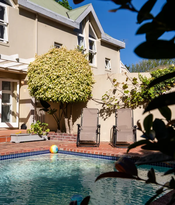 Sunny outdoor pool area with two lounge chairs, a colorful ball in the water, and a large manicured tree by the building.