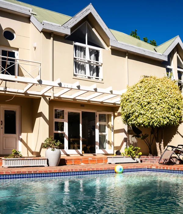 Sunny outdoor pool area with two lounge chairs, a colorful ball in the water, and a large manicured tree by the building.