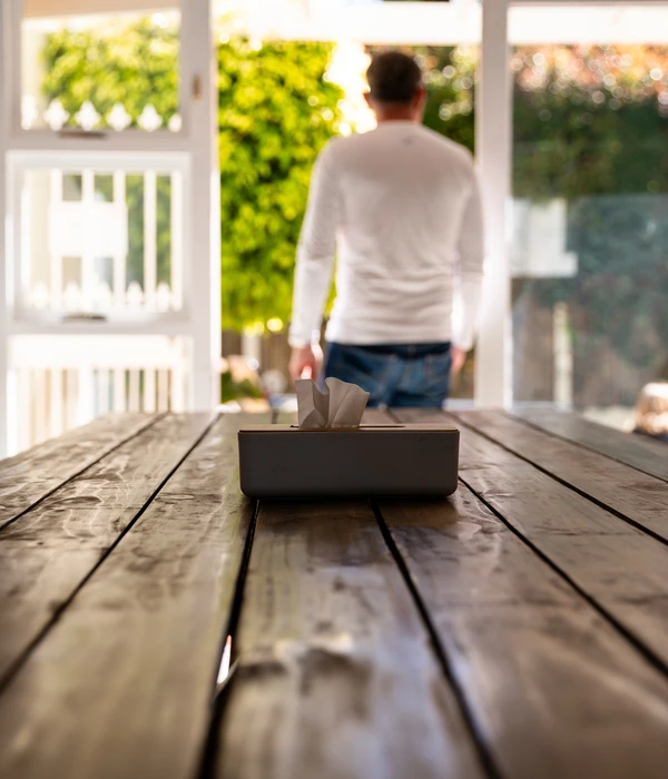 In focus, a tissue box on a long wooden table; out of focus, a man in a white shirt stands looking out a doorway.