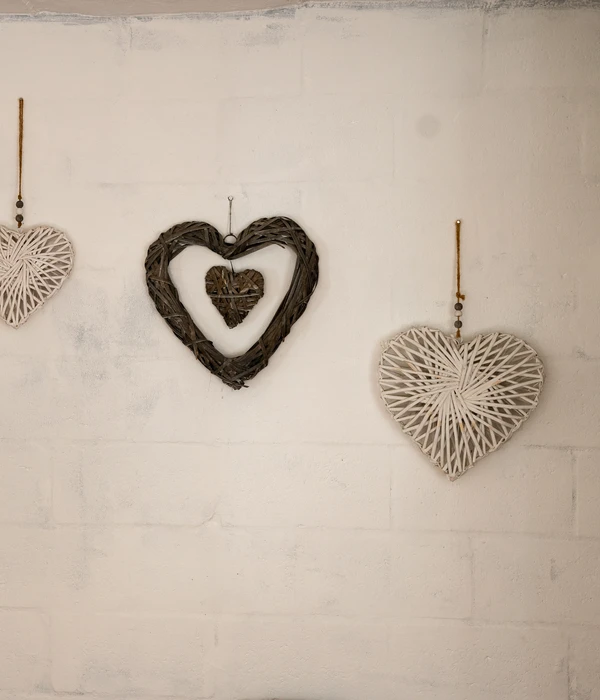 Three heart decorations hang on a white brick wall: a small white heart, a brown heart with a smaller one inside, and a large white heart.