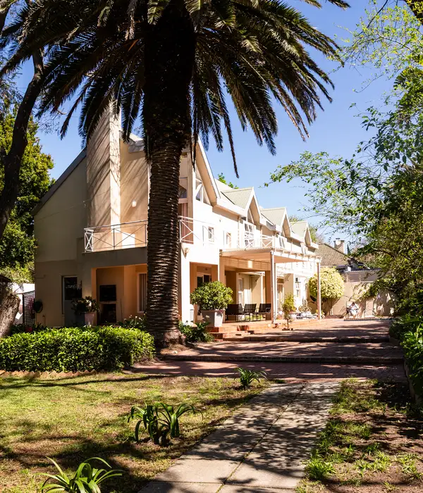 Exterior view of a large cream building with multiple gables, a balcony, a terrace with seating, and a tall palm tree.
