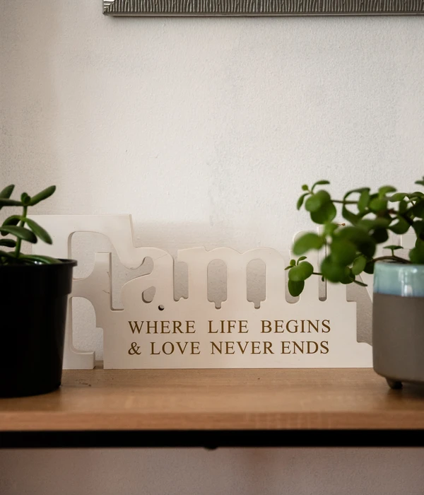 A white wooden "Family" sign on a shelf with the quote "Where life begins & love never ends" between two green potted plants.